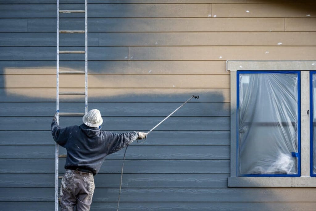 A professional painter uses a spray gun to apply exterior paint to a home's siding, with the window masked off for protection.