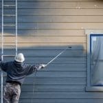 A professional painter uses a spray gun to apply exterior paint to a home's siding, with the window masked off for protection.